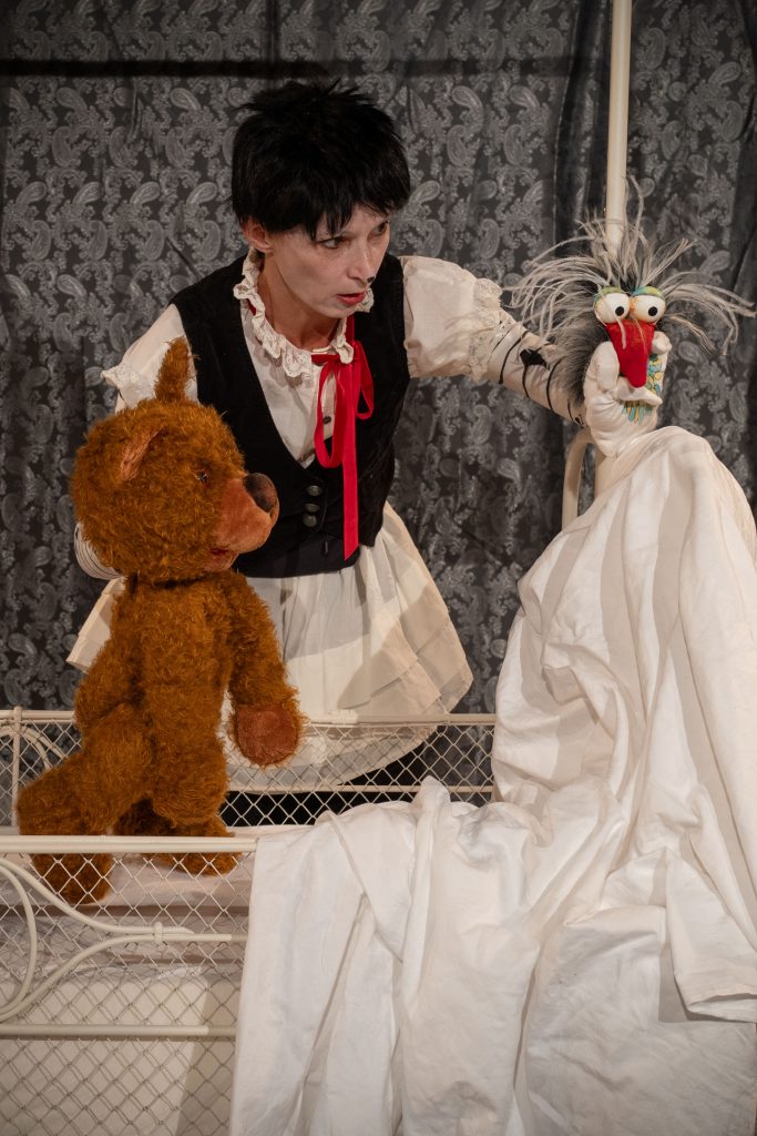 A person stands behind a white metal bed and operates two puppet figures. A brown bear puppet stands on the bed, while a bird puppet with large eyes and a red beak is surrounded by a white cloth. A gray patterned curtain forms the background.