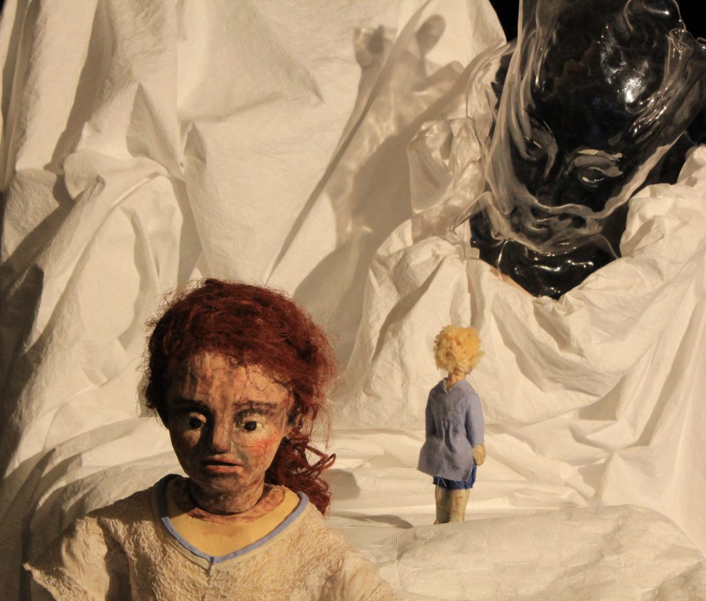 A puppet figure with curly red hair is positioned in the foreground against a backdrop of draped white fabric. A smaller figure with light hair and blue clothing stands in the background. A dark, mask-like object is partially visible in the upper right corner.