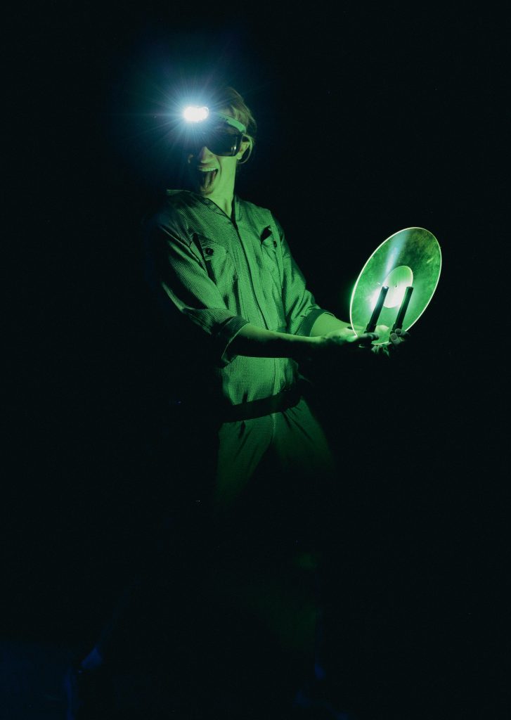 Eine Person steht in einem dunklen Raum und hält eine runde, reflektierende Scheibe in den Händen. Grünes Licht beleuchtet die Szene, während auf der Stirn eine helle Lichtquelle befestigt ist.