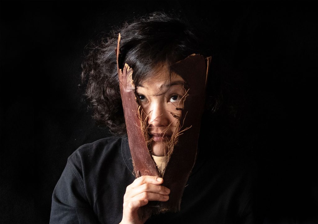 A close-up of a person’s face. The background and the person’s clothing are black. The woman is holding two pieces of tree bark in front of her face, forming the shape of the letter “V.”