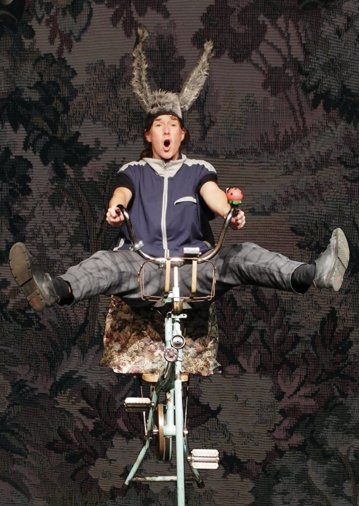 A person sits on a tall unicycle with arms and legs extended against a patterned stage backdrop. The person wears a costume with a headpiece featuring light feather-like elements. The unicycle is decorated with plant-like or natural materials.