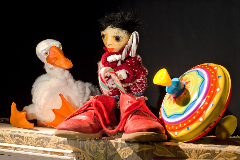 A figure with large eyes, a red sweater, and oversized red shoes sits among toys and props. Beside the figure are a white bird puppet with an orange beak and a colorful spinning top toy. The scene is illuminated against a dark background.