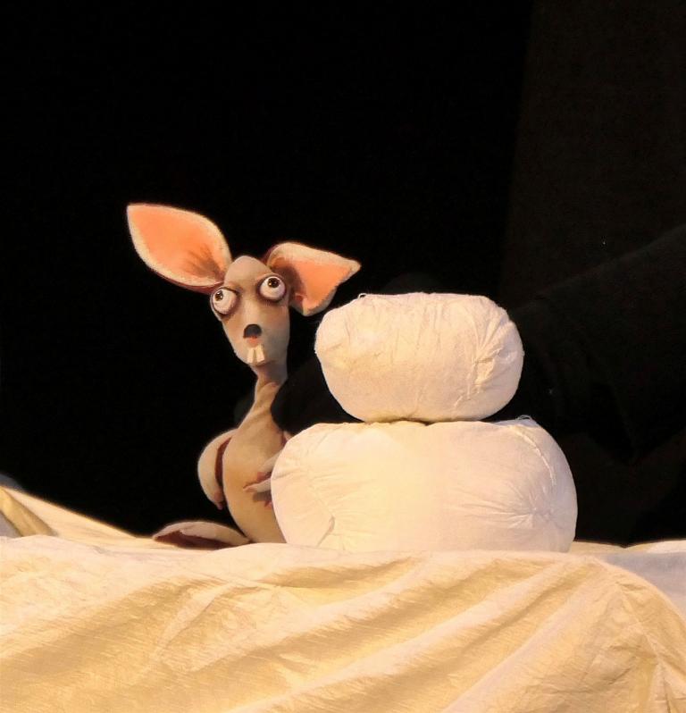 A small animal figure with large ears stands behind a stack of three white, cushion-like shapes. The scene is set against a dark background, with the light-colored surface and the white objects prominently highlighted in the foreground.