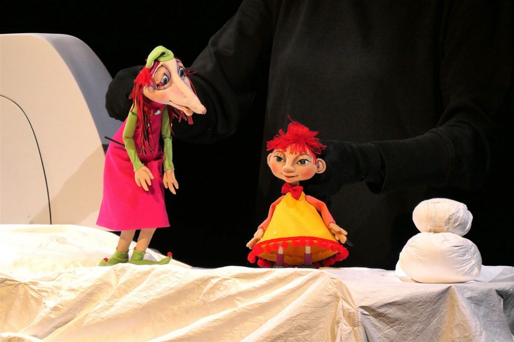 Zwei Puppenfiguren stehen auf einer hellen Stofffläche vor schwarzem Hintergrund. Eine Figur mit langem Kleid und grüner Kopfbedeckung steht links, während eine kleinere Figur mit rotem Haar und gelbem Kleid rechts daneben platziert ist. Die Szene ist theaterartig beleuchtet.