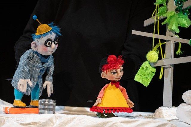 Three puppet figures stand on a light-colored fabric surface against a black background. A figure with red hair and a yellow dress is positioned in the center. On the left stands a blue-clad figure with long hair, while a green figure is partially visible on the right. The scene is lit like a theater stage.