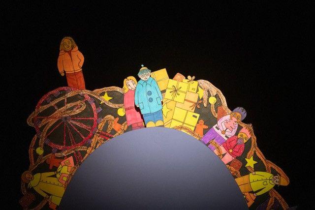 A row of colorful figures and houses is arranged on a semicircular platform against a black background. The elements are brightly colored and positioned along the upper edge of the curved structure.