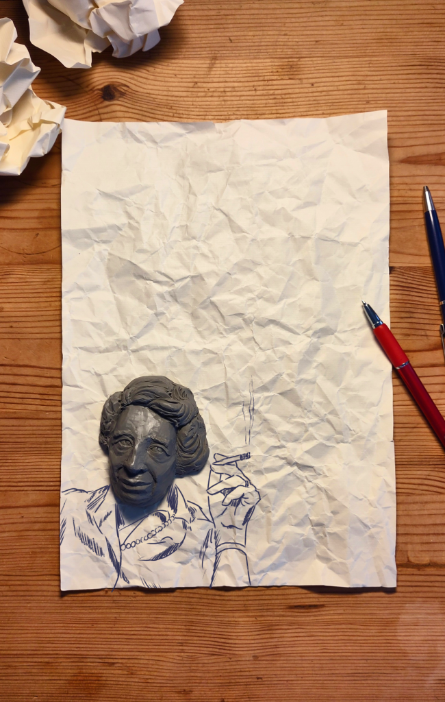 Auf einem Holztisch liegt ein zerknittertes Blatt Papier. Im unteren Bereich ist eine gezeichnete Person mit erhobener Hand und Zigarette zu sehen, deren Kopf als plastische graue Skulptur auf dem Papier platziert ist. Um das Blatt liegen mehrere zerknüllte Papierbälle sowie ein roter und ein blauer Kugelschreiber.