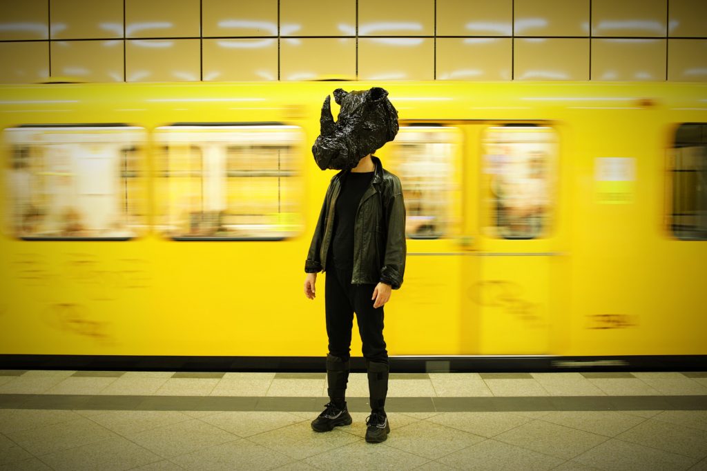 Person steht auf einem U-Bahn-Bahnsteig vor einem vorbeifahrenden gelben Zug. Die Person trägt dunkle Kleidung und hat den Kopf mit einem dunklen, wolkenartigen Objekt verdeckt. Der Hintergrund ist durch die Bewegung des Zuges leicht verschwommen.