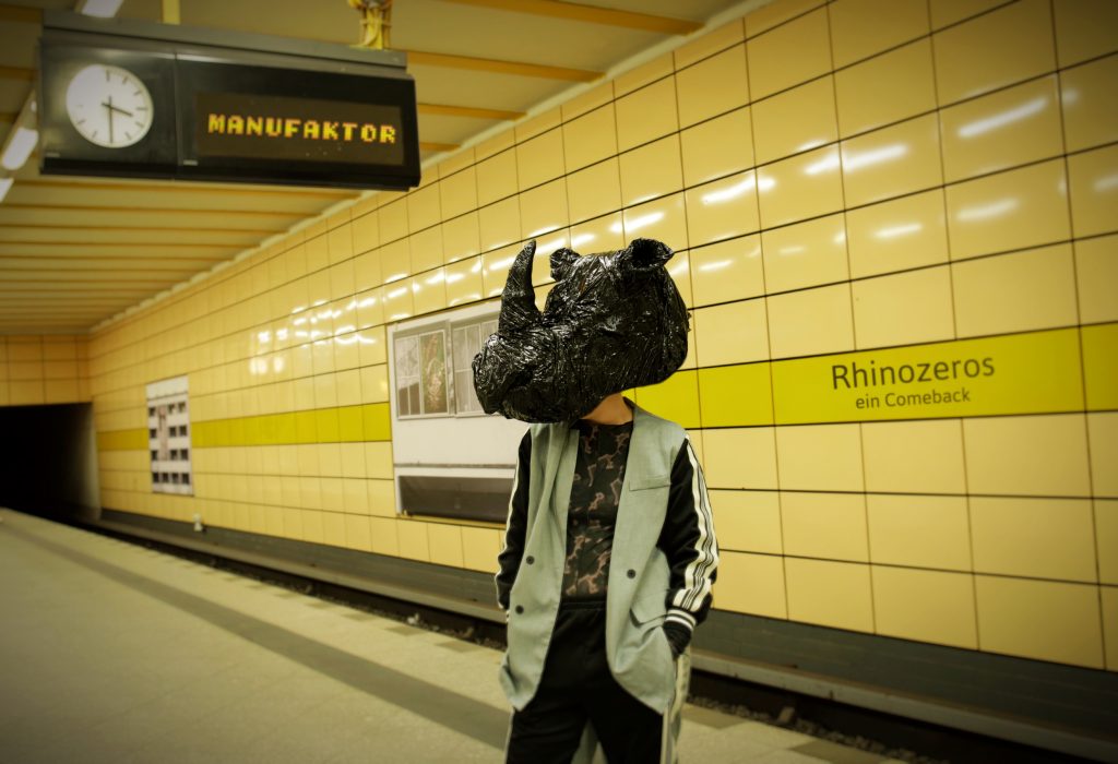 English translation: A person is standing on a subway platform in front of a passing yellow train. The person is dressed in dark clothing, and their head is obscured by a dark, cloud-like object. In the background, a sign reading "Rhinozeros ein Comeback" hangs in place of the station name.