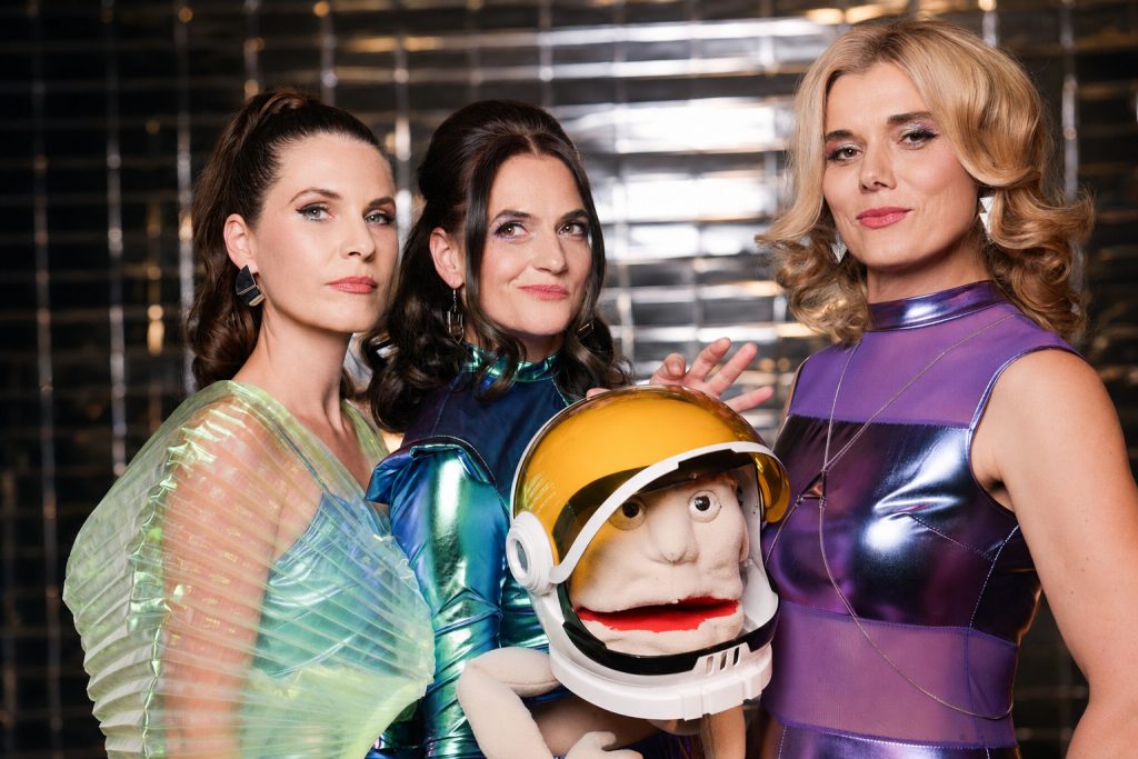 Drei Personen in glänzenden, futuristisch wirkenden Kostümen posieren vor einem reflektierenden Hintergrund. In der Mitte ist eine Handpuppe mit großem Astronautenhelm. Die Kostüme schimmern in Grün-, Blau- und Lilatönen.