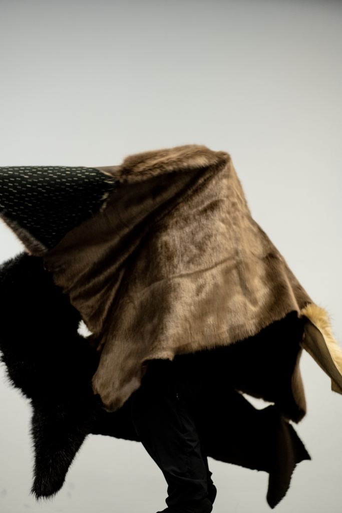 Brown fabric draped over a black, fur-covered sculptural form against a light background. The image highlights the contrast between the soft folds of the fabric and the textured surface of the fur.
