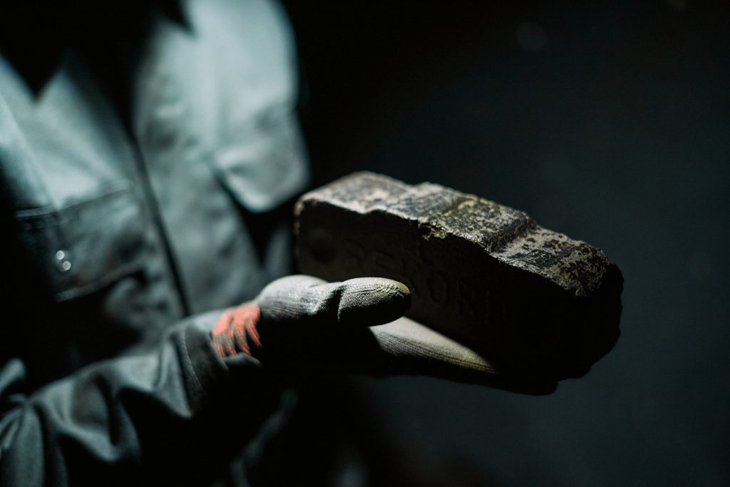 Eine Person in Arbeitskleidung und Schutzhandschuh hält einen dunklen Ziegelstein oder Gesteinsblock in einer ausgestreckten Hand. Die Aufnahme ist stark fokussiert auf das Objekt im Vordergrund, während die Person und der Hintergrund im Schatten liegen.