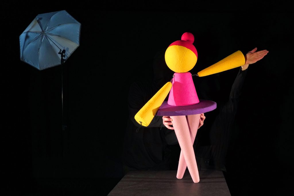 Abstract puppet figure in yellow and pink on a stage against a black background.