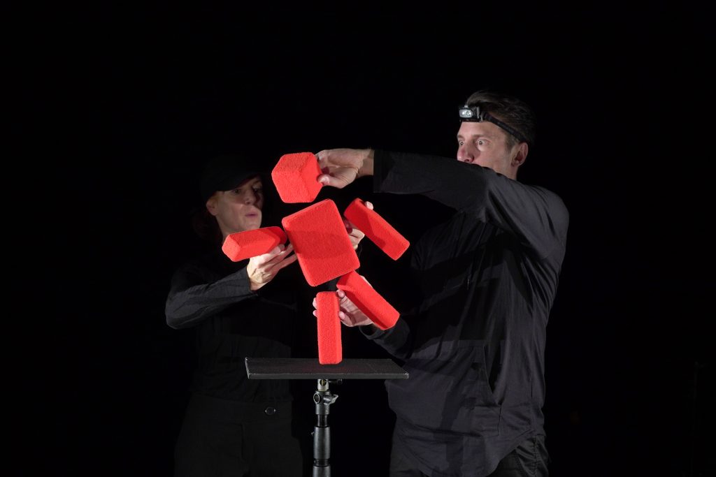 Abstract red puppet figure made of geometric shapes, manipulated by two puppeteers.