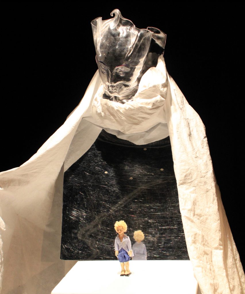 Two small figures stand in front of a large dark stage or backdrop. A light-colored fabric is draped along the sides, framing the scene. The figures are positioned near the bottom of the image, while the tall backdrop occupies most of the composition.