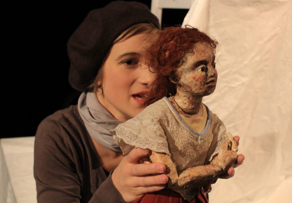 A person holds a small puppet figure with curly red hair and a light-colored dress in front of a light fabric backdrop. The puppet is seated with bent arms while being supported by the person's hands. The scene is warmly lit and shown in close-up.