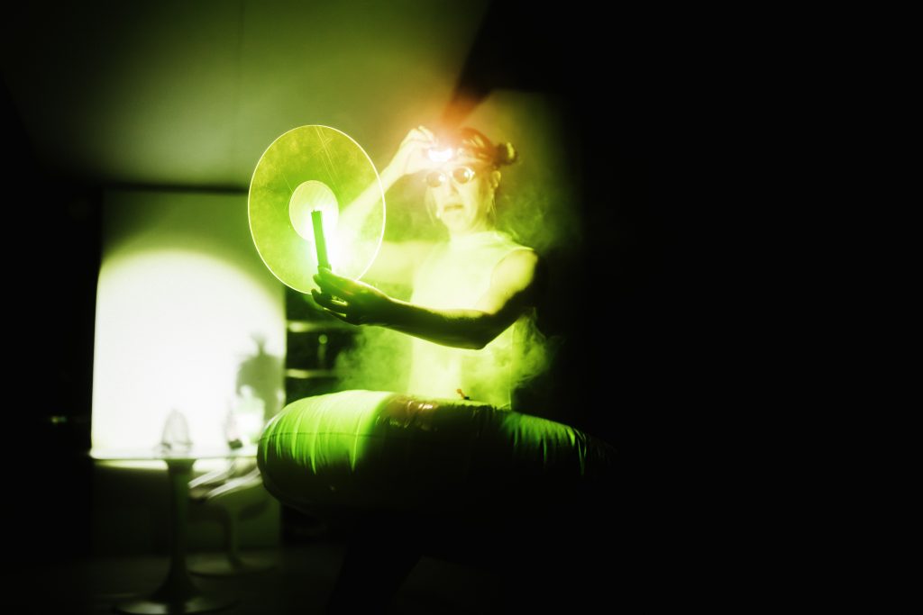 Person in a dark setting surrounded by green light and mist, holding a large transparent circular disc.