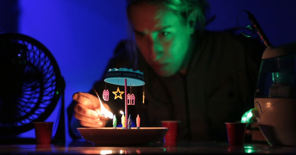A person lights candles on a small birthday cake. The table is illuminated with blue and green lighting. A fan, several red cups, and a humidifier are positioned beside the cake.