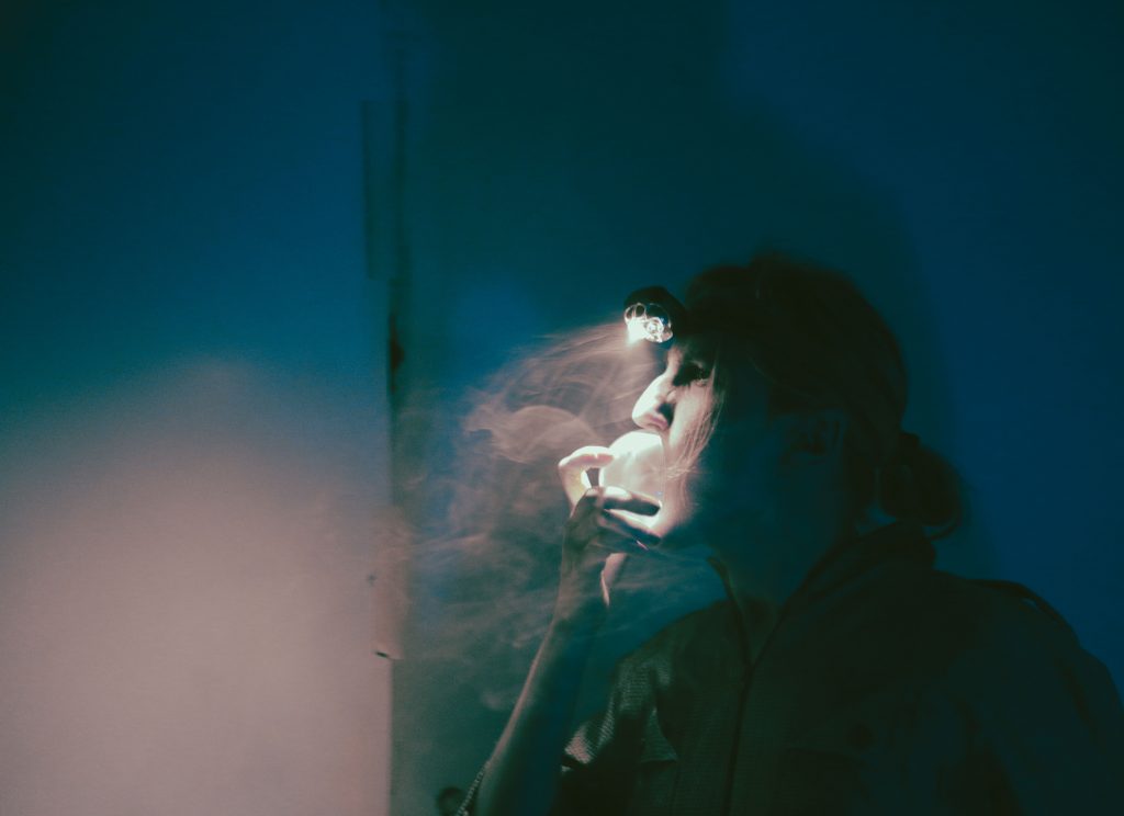 A person is shown in partial profile against a dark background. A visible cloud of vapor or mist emerges from the mouth. Blue and green lighting illuminates the scene, highlighting the face and upper body.