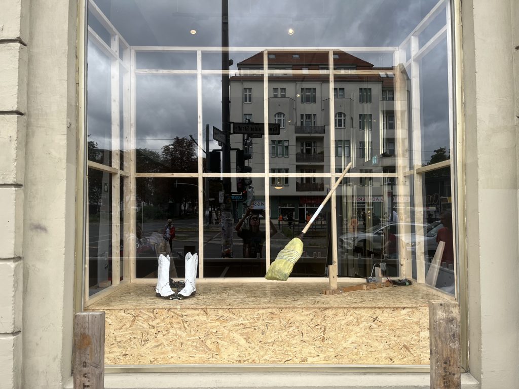 Schaufenster mit Holzgerüst und Holzplattform. Auf der Plattform stehen weiße Stiefel, daneben ist ein Besen befestigt. In der Fensterscheibe spiegeln sich Straße und Gebäude.