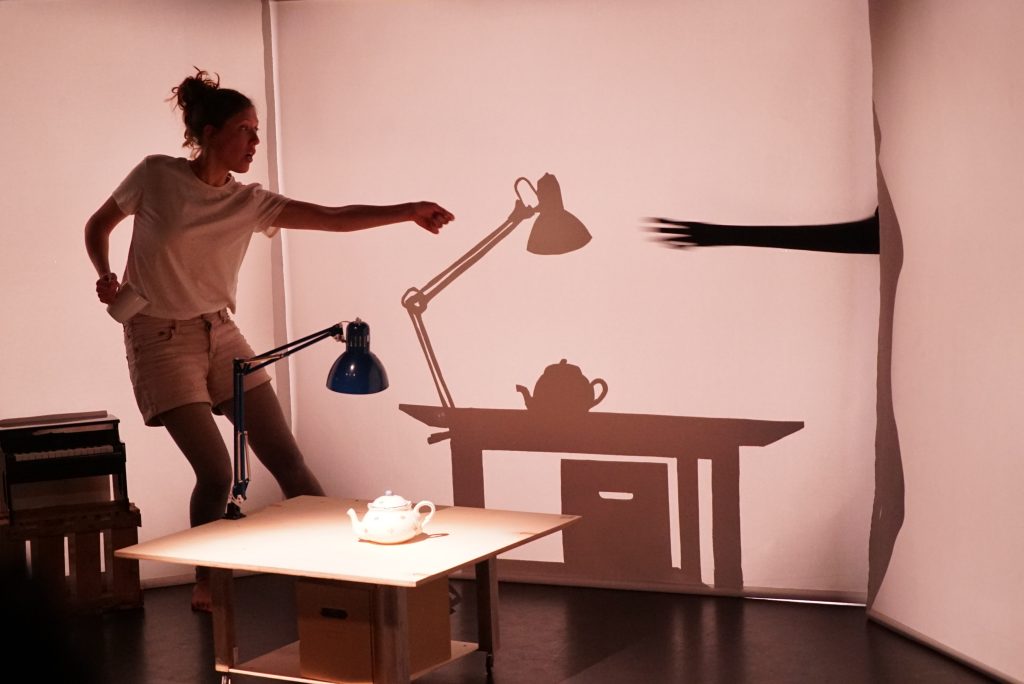 Person erzeugt mit Licht und Schatten eine Szene aus Tisch, Lampe und Teekanne.