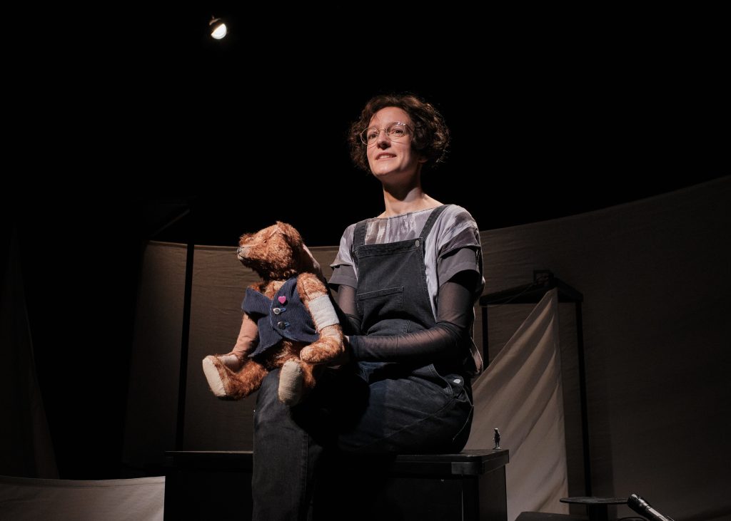 Person sitzt mit einer Handpuppe (Bär) auf einer Theaterbühne.