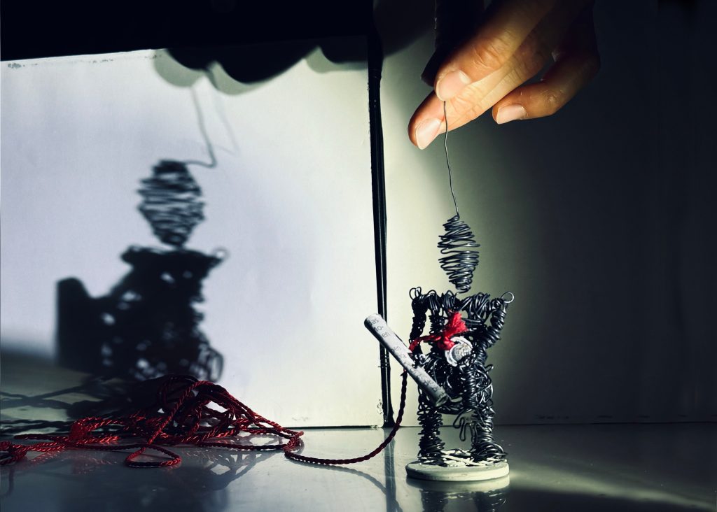 hand guides a wire figure with a red string before a lit backdrop.