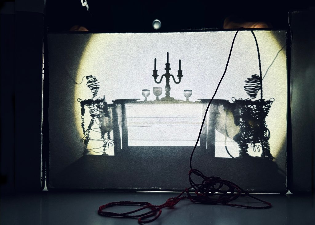 Shadow play with two wire figures at a set table with a candelabra.