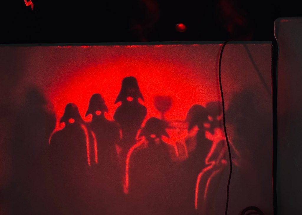 Rotes Schattenspiel mit silhouettierten Figuren vor dramatischem Hintergrund.
