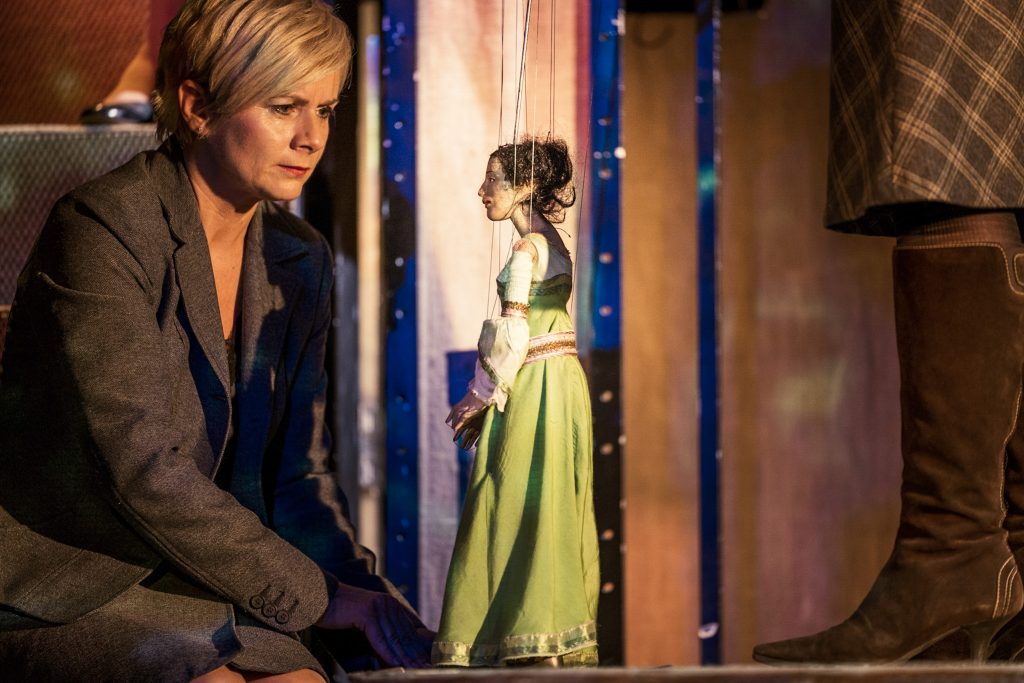 Figurenspieler:in neben einer Marionette im grünen Kleid auf einer Theaterbühne.