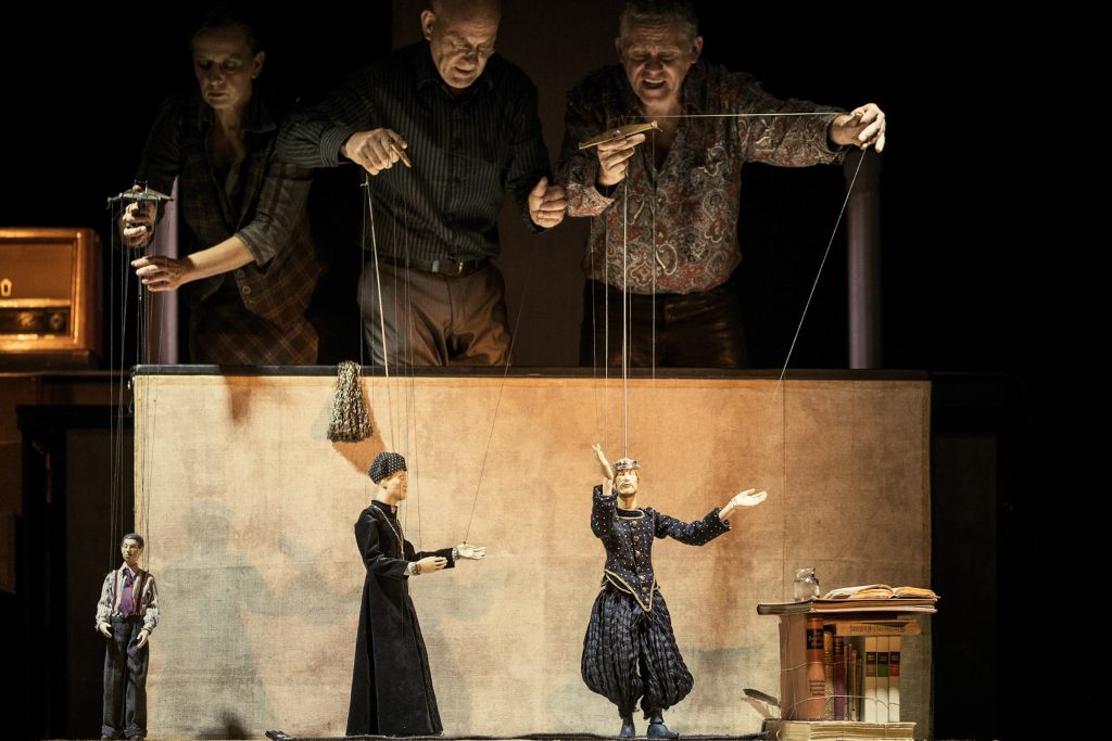 Three puppeteers operate puppet figures on a stage with a table setting.