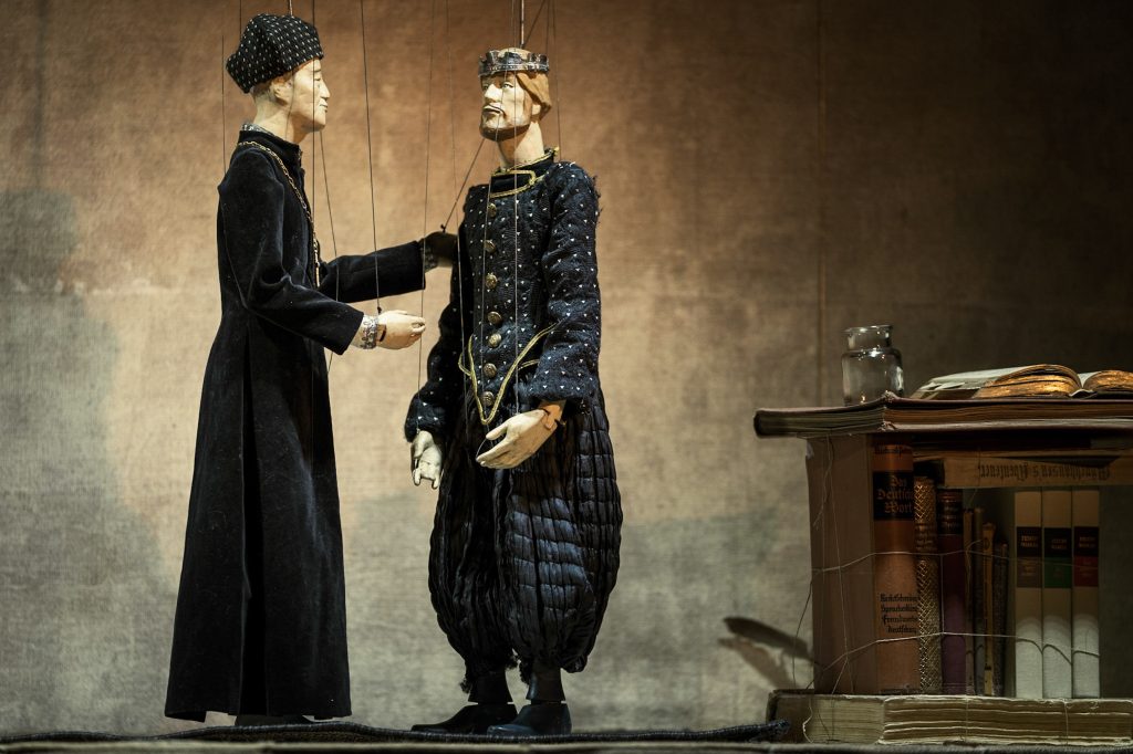 Two puppet figures in dark costumes stand on a stage beside a table.